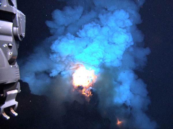 The eruption produces a bright flash of hot magma that is blown up into the water before settling back to the seafloor. In the foreground is the front of the Jason remotely-operated vehicle (ROV) with sampling hoses. The area in view is about 6-10 feet across. 
