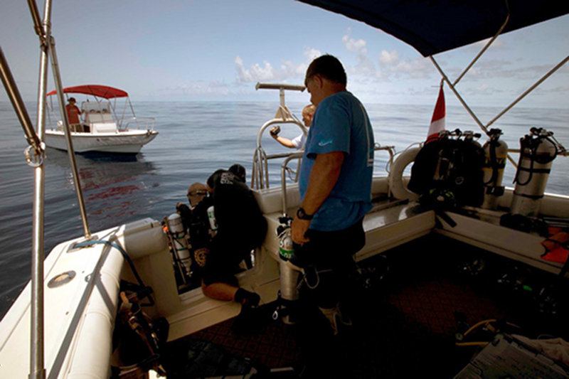 Brett Gonzalez assists Tom Iliffe with his bailout tanks as he prepares to dive on a beautiful flat sea. Alex Chequer stand by in a fast boat in case of emergency. Project DSO, Brian Kakuk, watches over the operation.