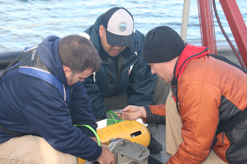 Evan Martizal, Vitad Pradith, and Matthew Lawrence ready the bow­mounted swath bathymetry and side­scan sonar sensor for deployment.