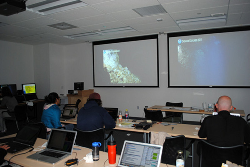 Team Rhode Island in the Exploration Command Center watching video streaming from NOAA Ship Okeanos Explorer at the Mid-Cayman Rise. The console and intercom system is on the left of the image and scientists can also communicate with each other and the ship by instant messaging from computers.