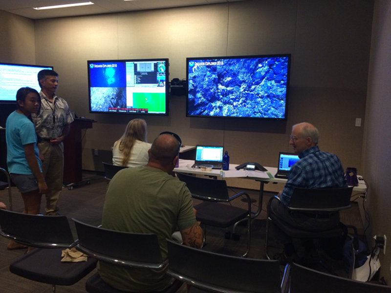 Bruce Mundy (far right), Fish Biologist at NOAA Pacific Island Fisheries Science Center, discussing fish with a group of NOAA scientists and visitors with the live-video feed in the background at the NOAA Inouye Regional Center.