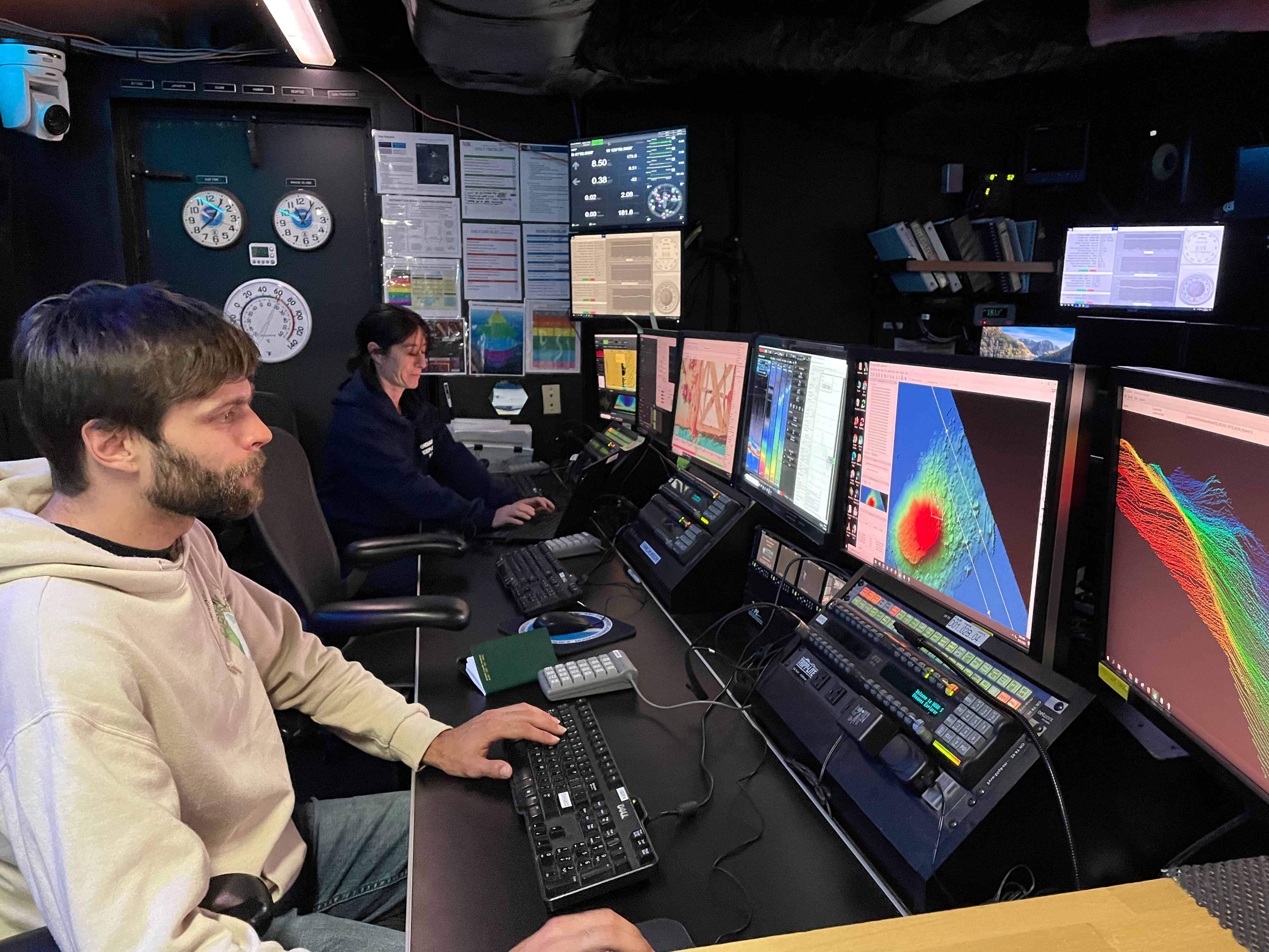 Two explorers-in-training working on computers in the control room of NOAA Ship Okeanos Explorer. One works on the map of a seamount in the Pacific Ocean another supervises data collection.