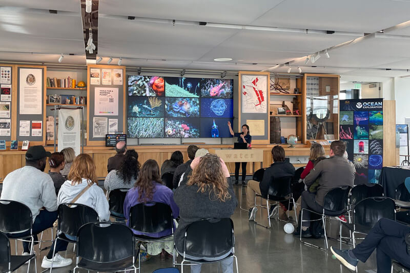 An audience listening to a speaker with images from the deep ocean on a screen behind her.