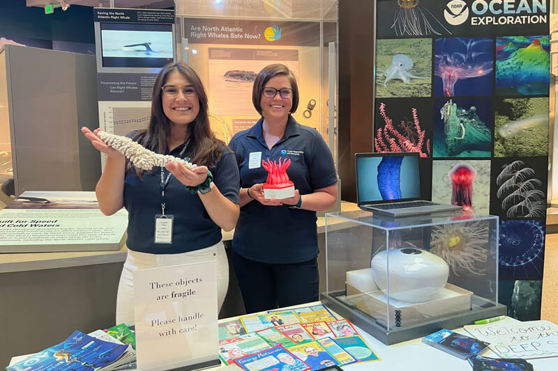 Two women standing by a table with ocean-exploration related materials in a museum.