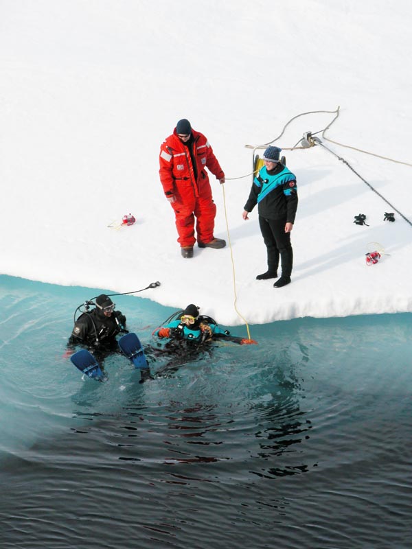 Ice Divers enter the water while a Coast Guard Petty Officer tends the line and a safety diver stands by.