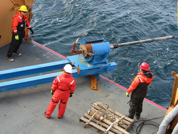 The gravity corer relies on a weight at the top to drive the barrel into the sediment. In this photo, the weight, with a 10-ft barrel attached, is cradled in the blue 'bucket', which has been moved to the end of a specially built track. The bucket will be rotated 90° so that the core barrel points straight down, then the corer will be winched out of the bucket, into the water, and down to the seafloor. Standing by (right to left) are Pete, Jenny, and Coast Guard Marine Science Technician MST3 Marshal Chaidez.