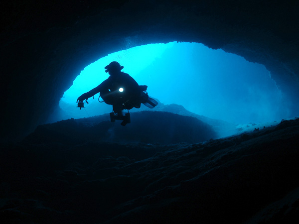 A diver entering a pitch black cave is silhouetted by sunlight filtering through the depths above.