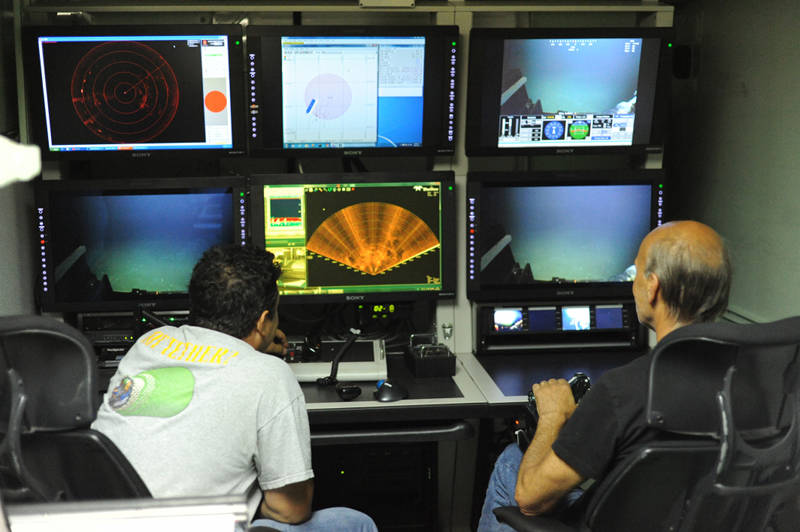 Figure 1: Jamie (right) and Tony (left) at the controls of the ROV Global Explorer. In Jamie’s left hand is the joystick that he uses to pilot the vehicle. The various monitors in front of them tell them what the ROV sees and where it is. Tony is monitoring the SONAR (lower middle monitor) to find a deep-sea camera system.