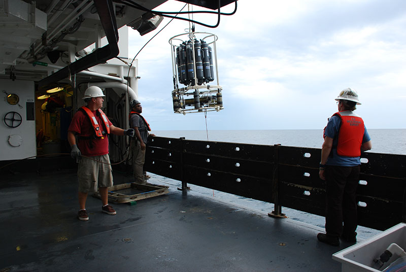 Amanda Demopoulos watches as deck crew members Knott and Cornell deploy the CTD at Pea Island B. The CTD collects conductivity (salinity), temperature, and depth (pressure) data, plus it has sensors to detect dissolved oxygen, chlorophyll, and turbidity (particles in the water), as the instrument travels down to the seafloor. The CTD is also equipped with a rosette of Niskin bottles, which are specially designed water bottles that can be triggered to collect water at set depths.