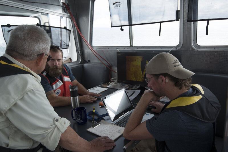 Dr. Guy Meadows (left) and Chris Pinnow (right) of Michigan Technological University discuss sonar imagery with Thunder Bay National Marine Sanctuary Research Coordinator John Bright (center). The image shown on the computer screen is from the site believed to be Choctaw. Acoustic data was collected via an autonomous underwater vehicle (AUV) operated by MTU during field operations in June, 2017.