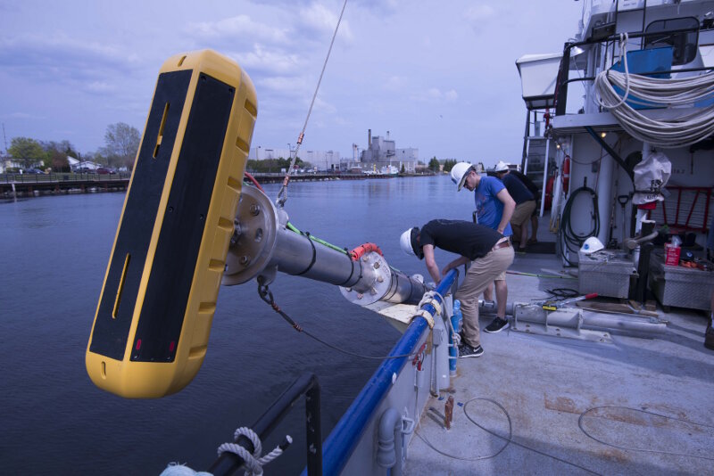University of Delaware sonar technicians Kenny Haulsee and Peter Barron route cables from the echosounder (yellow device in foreground) along a pole mount used to lower and secure the echosounder along the side of the vessel.