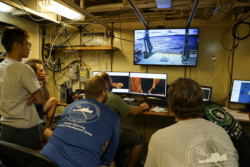 On board Research Vessel Kilo Moana during the Deepwater Surveys of World War II U.S. Cultural Assets in the Saipan Channel expedition, researchers from Scripps Institution of Oceanography examine side-scan sonar data (visible on screen in a bronze color) for anomalies that could indicate aircraft wreckage. Side-scan sonar data were collected from a REMUS 600 in water depths as great as 600 meters (1,967 feet). The data reveal a detailed picture of the seafloor, including measurements of the size of objects and how high objects project above the seafloor. The large monitor shows the researchers a real-time view of the deck of the ship, where preparations for deploying a remotely operated vehicle are taking place.