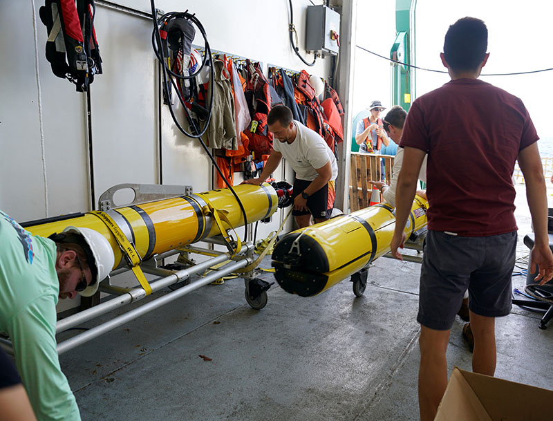 The team prepares the REMUS 600 autonomous underwater vehicles for launch in the Saipan Channel as part of the Deepwater Surveys of World War II U.S. Cultural Assets in the Saipan Channel expedition. Each REMUS vehicle is equipped with side-scan sonar, multibeam sonar, and a low-light imaging camera. This view is looking out of the loading bay of Research Vessel Kilo Moana, where the vehicles can undergo maintenance between dives.