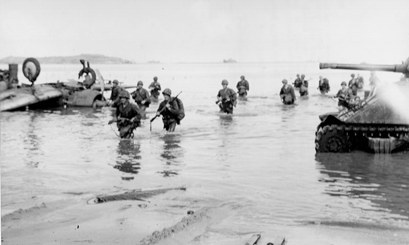 Marines wading ashore to the beach at Guam, while the bombardment went on overhead. The tank covered their landing as they walked through water past a Japanese plane downed while attempting to thwart the landing operations.