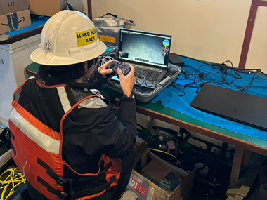 The image depicts a person seated at a workbench, operating a laptop with a gaming controller. The individual is wearing a white hard hat labeled "HARD HAT AREA" and a high visibility safety vest with orange and black colors. The person is focused on the laptop screen displaying a camera feed.