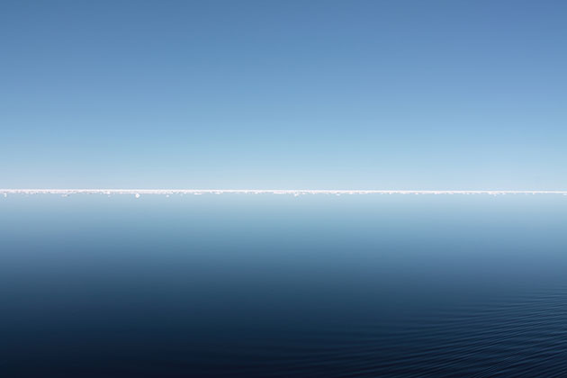 A stunningly blue and calm Arctic reflection of sea and sky divided by distant bright white ice and interrupted by ripples created by the ship. 15th June 2012 on the RRS James Clark Ross in the Arctic sea ice between Svalbard and Greenland. 