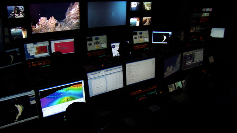   View of the control room on board <em>Okeanos Explorer</em> during ROV operations. You can see live footage from the ROV on the left monitor, revealing sulphur chimneys covered with dense communities of barnacles on the seafloor.