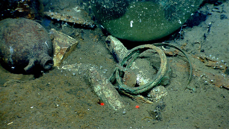 Two depth sounding leads used to measure the depth of water under the vessel and to characterize the local seafloor and a brass frame and gimbal band, likely for a compass, all nestled among glass and ceramic bottles and jugs.