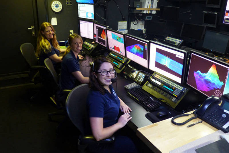Collecting and processing high-resolution mapping data is done from the Mission Control room. Here we monitor the sonars, clean and process files, and use specialized software to visualize seafloor features in three-dimensions. From left to right: Kasey Cantwell, Field Operations Specialist with NOAA Office of Ocean Exploration and Research; Lindsay McKenna, Expedition Coordinator with NOAA Office of Ocean Exploration and Research; and Abigail Casavant, Explorer-in-Training from the University of Rhode Island History and Underwater Archeology graduate program.