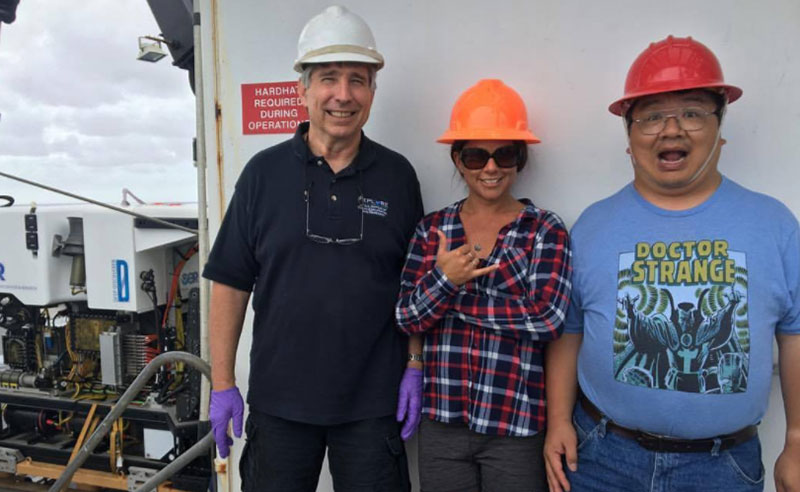 Expedition science leads, Dr. Chris Kelley and Dr. Chris Mah, and NOAA EPP Intern, Nikola Rodriguez, pose for a picture while standing by waiting to receive word that the ROV is secured on deck and it is safe for them to go retrieve samples collected during the day’s ROV dive.