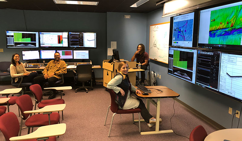 Image of the NOAA Ocean Exploration onshore mapping team at the University of New Hampshire Exploration Command Center participating through telepresence during the first part of the Windows to the Deep 2018 expedition. This image includes three of the explorers-in-training who participated in this expedition from shore.