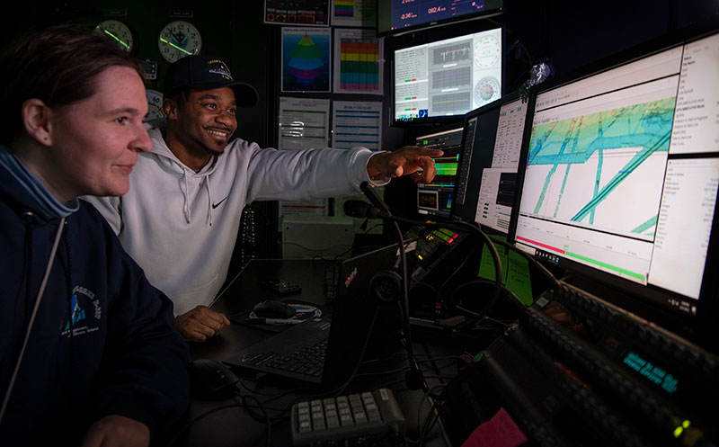Explorers-in-training Daryin Medley and Kathrin Bayer in the control room of NOAA Ship Okeanos Explorer viewing multibeam bathymetry data collected during the 2022 Caribbean Mapping expedition.