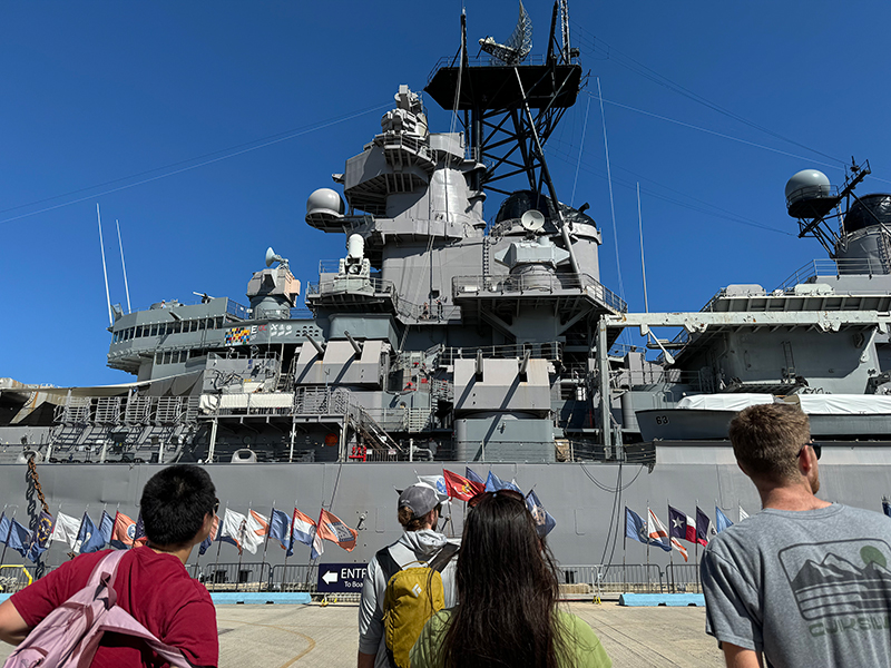 Image of explorer-in-training Elizabeth, media specialist Nate, explorer-in-training Anabel, and cultural liaison Makoa looking up at the giant USS Missouri.