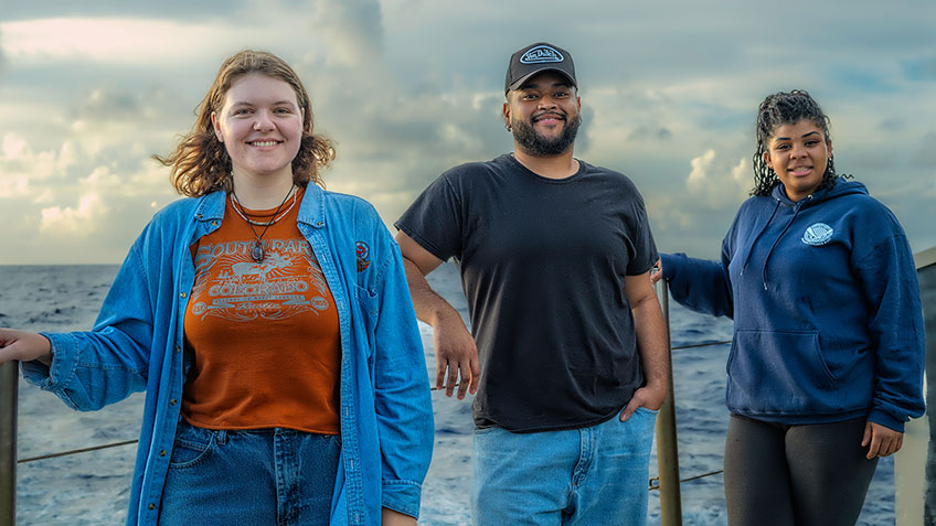 Three people standing on a vessel, with the sea and cloudy sky in the background.