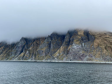 Steep cliffs and dense clouds bid the ship farewell on departure from Unalaska.