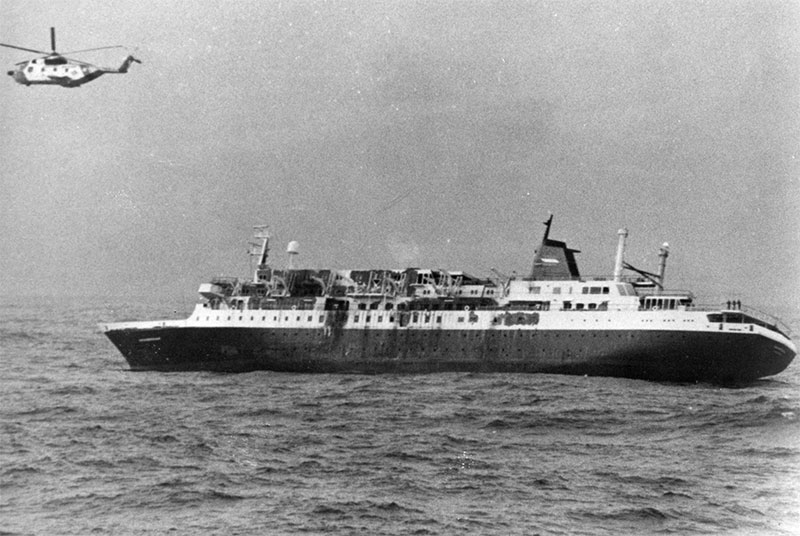 Black and white historic photo of a helicopter flying above a cruise liner starting to turn on its side.