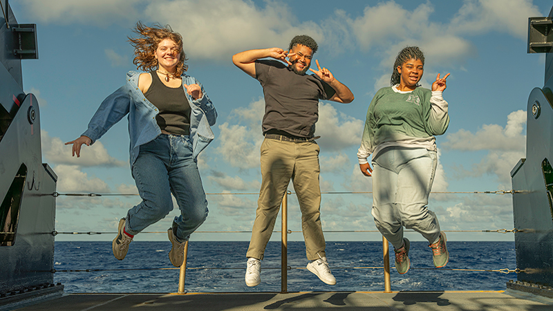 Three people jumping on a ship's deck with the ocean and sky in the background.